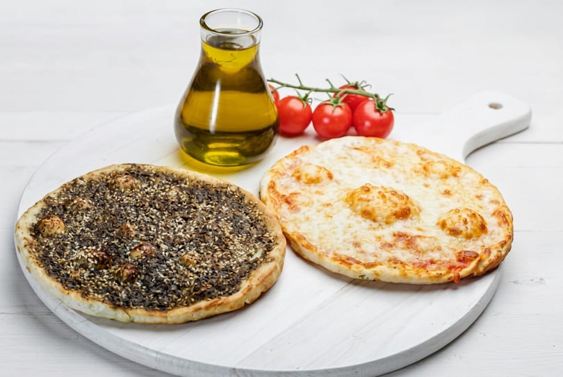 Two round flatbreads, one with za'atar and one with cheese, next to olive oil and tomatoes.