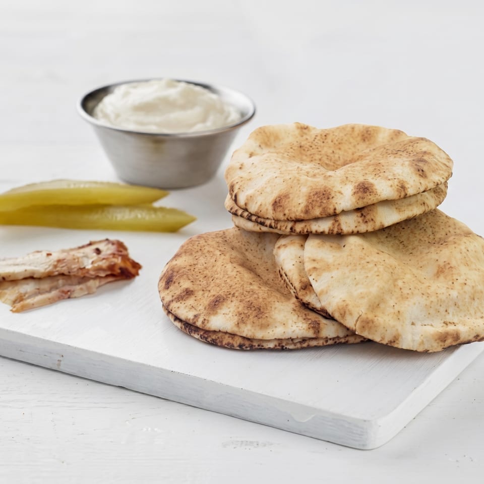Pita bread, pickles, chicken, and a bowl of white sauce on a white wooden board.