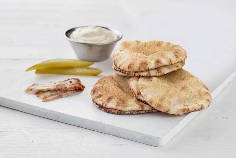 Pita bread, pickles, chicken, and a bowl of white sauce on a white wooden board.