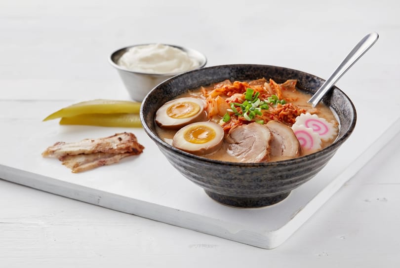 A bowl of ramen with pork, eggs, kimchi, and green onions on a white cutting board.